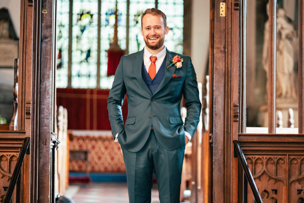 A smiling man in a blue suit with an orange tie and boutonnière stands with his hands in his pockets inside a church, with stained glass windows in the background.