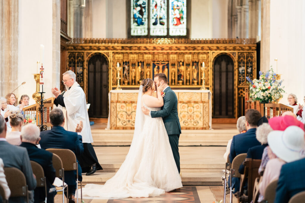 A bride and groom share their first kiss at the altar during a church wedding ceremony, with guests seated and a priest standing nearby. The altar and stained glass windows are visible in the background.
