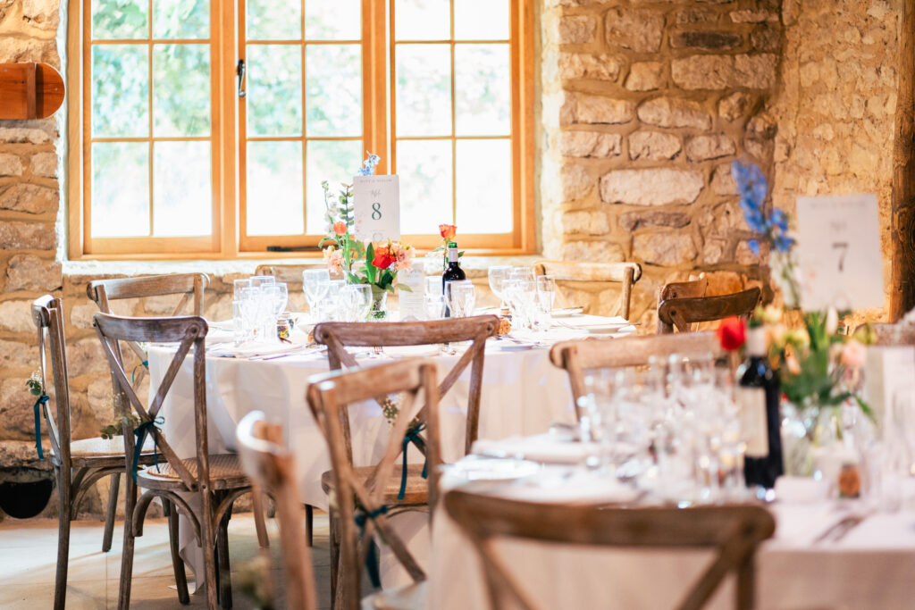 A rustic dining room with stone walls and wooden chairs, featuring round tables set with white tablecloths, glassware, floral centerpieces, and numbered table signs near a sunlit window.