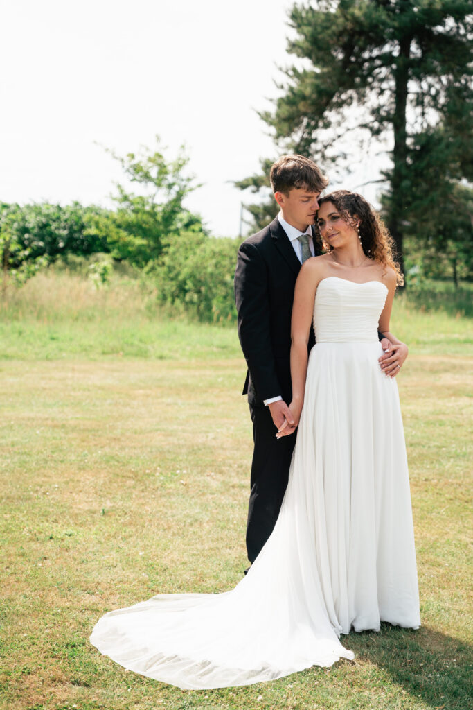 A bride in a strapless white gown and a groom in a black suit stand together on a grassy lawn, holding hands and looking at each other with trees in the background.
