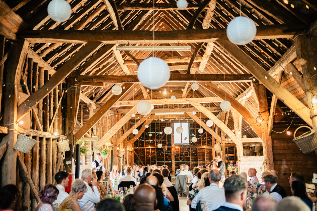 A large group of people are seated at tables inside a rustic wooden barn decorated with white paper lanterns and string lights, attending a festive event or celebration.