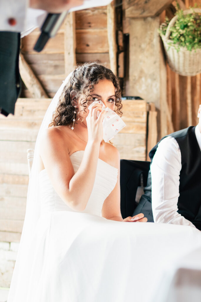 A bride in a white dress and veil sits indoors, wiping her eye with a tissue, appearing emotional during a wedding event. A person in a suit is seated next to her. The background is rustic wood.