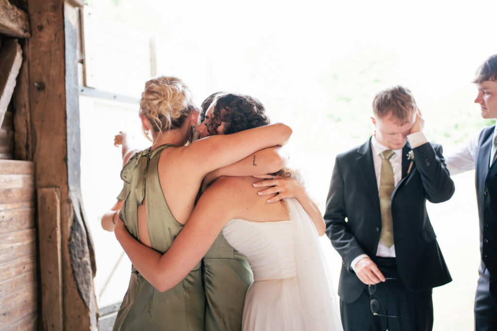 Two women, one in a white dress and one in green, hug closely at an indoor event. Nearby, two men in suits stand, one wiping his face, creating an emotional, heartfelt scene.