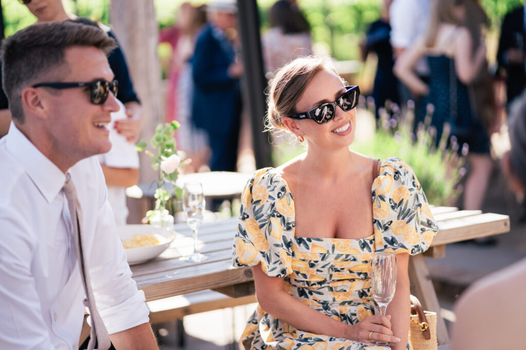 A woman in a yellow floral dress and sunglasses smiles while holding an empty wine glass, sitting beside a man in a white shirt and sunglasses at an outdoor social gathering. People mingle in the background.
