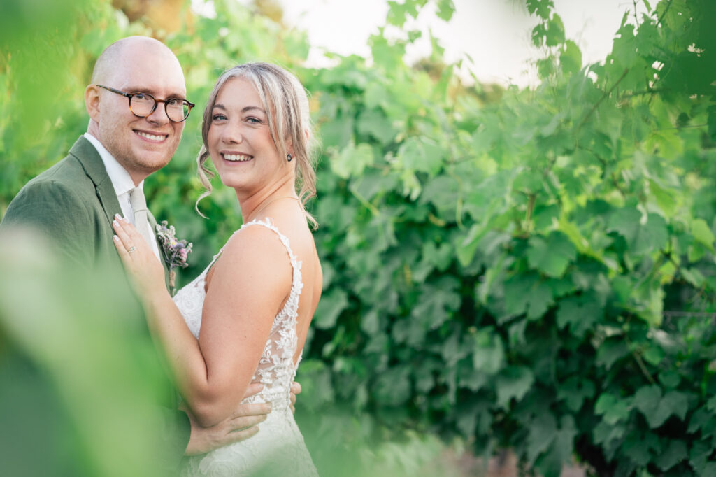 A smiling bride and groom stand close together outdoors, surrounded by lush green foliage. The bride wears a lace wedding dress, and the groom is in a green suit with glasses. Both look happily at the camera.