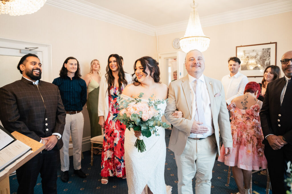 A bride holding a bouquet smiles while walking down the aisle with an older man, likely her father. Guests stand around them, smiling and watching in a bright room with chandeliers.