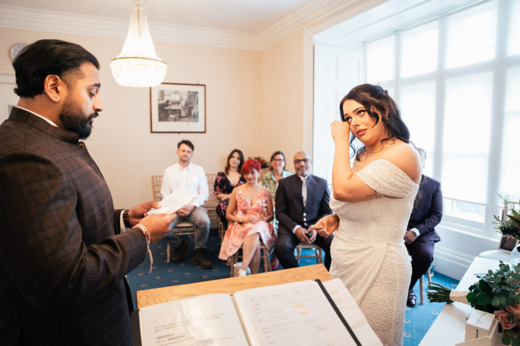 A bride in a white dress wipes a tear as the groom reads vows in a small room. Seated guests watch the ceremony, with sunlight streaming through large windows in the background. A document is visible in the foreground.