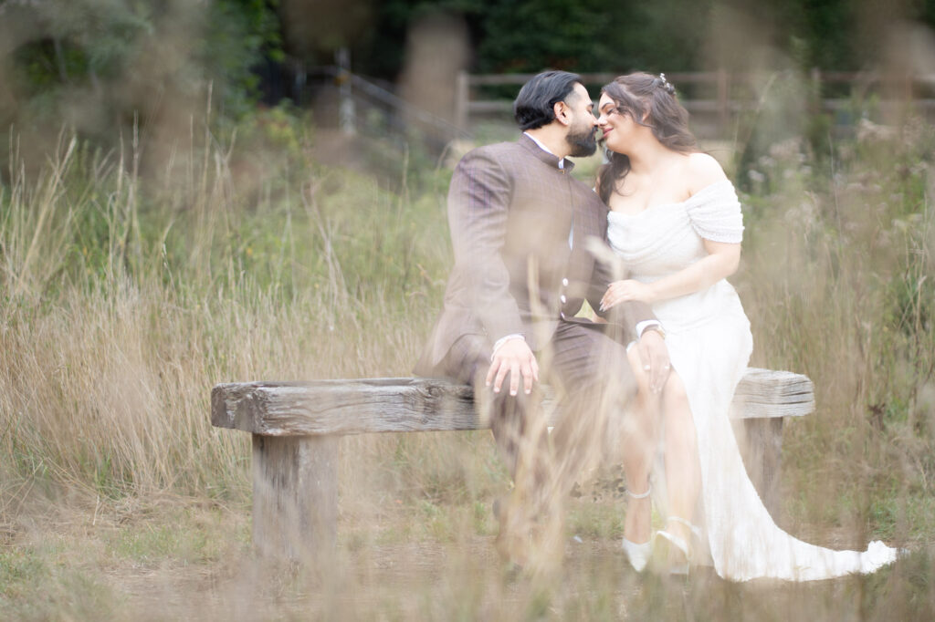A couple dressed in formal attire sits closely on a wooden bench outdoors, leaning in for a kiss. The woman wears a white dress, and the scene is framed by tall grass with a blurred natural background.