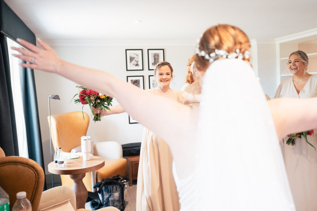 A bride with outstretched arms, seen from behind, greets two smiling bridesmaids holding bouquets in a bright, cozy room with modern decor and framed photos on the wall.