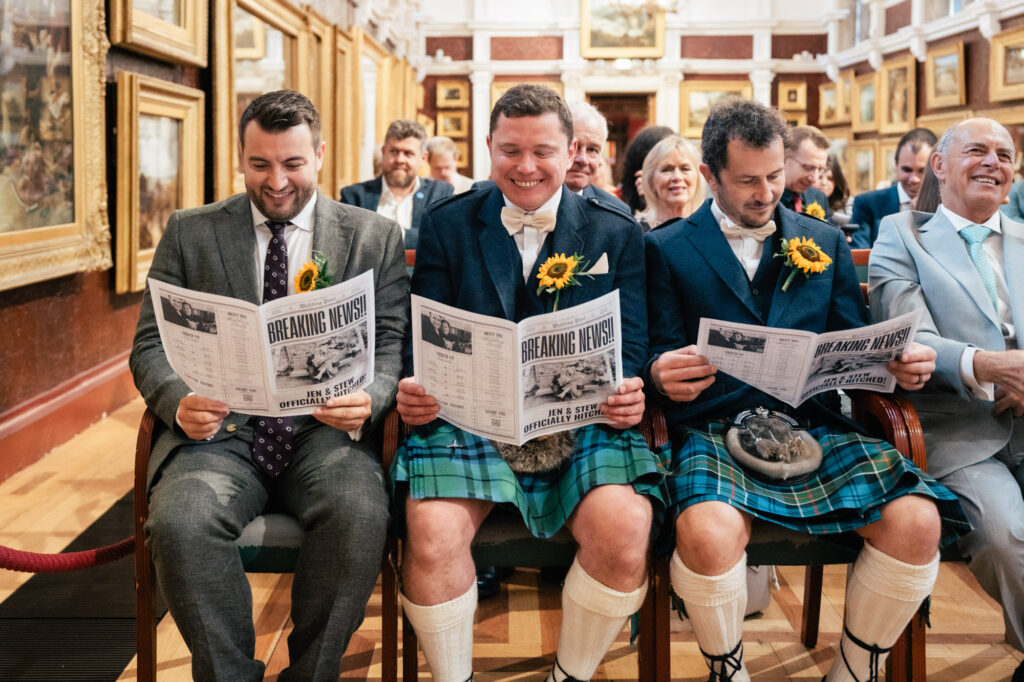 Three men, two in kilts and one in a suit, sit smiling while reading wedding programs designed like newspapers. The setting is an art gallery, and other guests are seated around them, all looking cheerful.