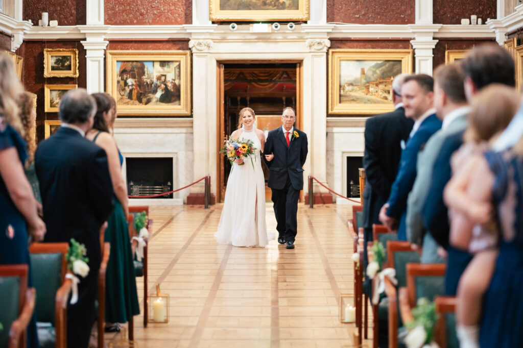 A bride in a white dress holding a bouquet walks down the aisle with an older man in a suit, while guests seated on either side watch in a grand room decorated with paintings.