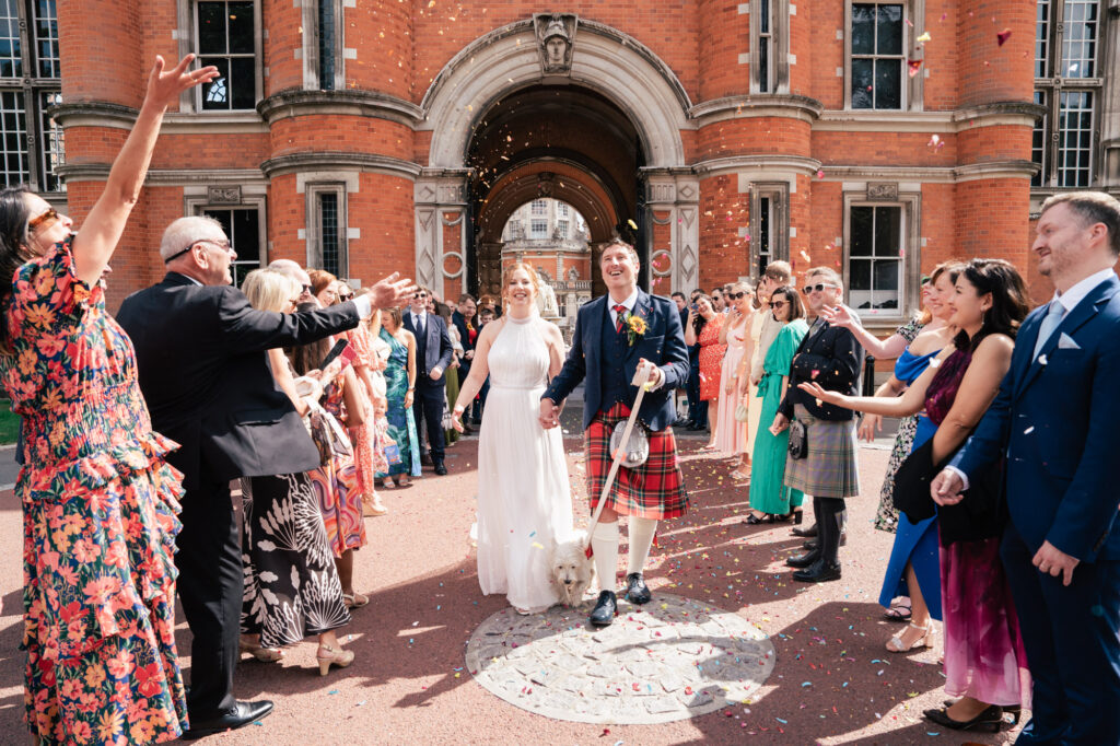 A newlywed couple, with the groom in a kilt and the bride in a white dress, walk hand-in-hand outside a red-brick building as guests cheer and throw confetti. A small dog walks beside them.