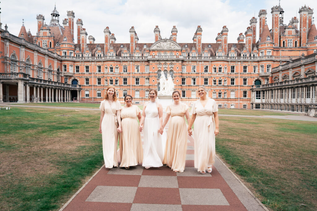 Five women in matching pale dresses walk together on a checkered pathway in front of an ornate, historic red-brick building with many windows and towers, on a cloudy day.