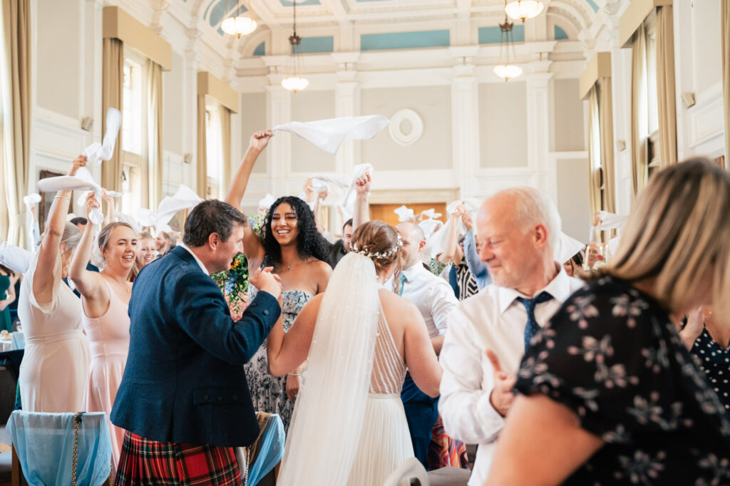 A joyful wedding reception scene in a bright hall, with guests standing, waving napkins in the air, and smiling. The bride and groom face each other in the center, surrounded by cheering friends and family.