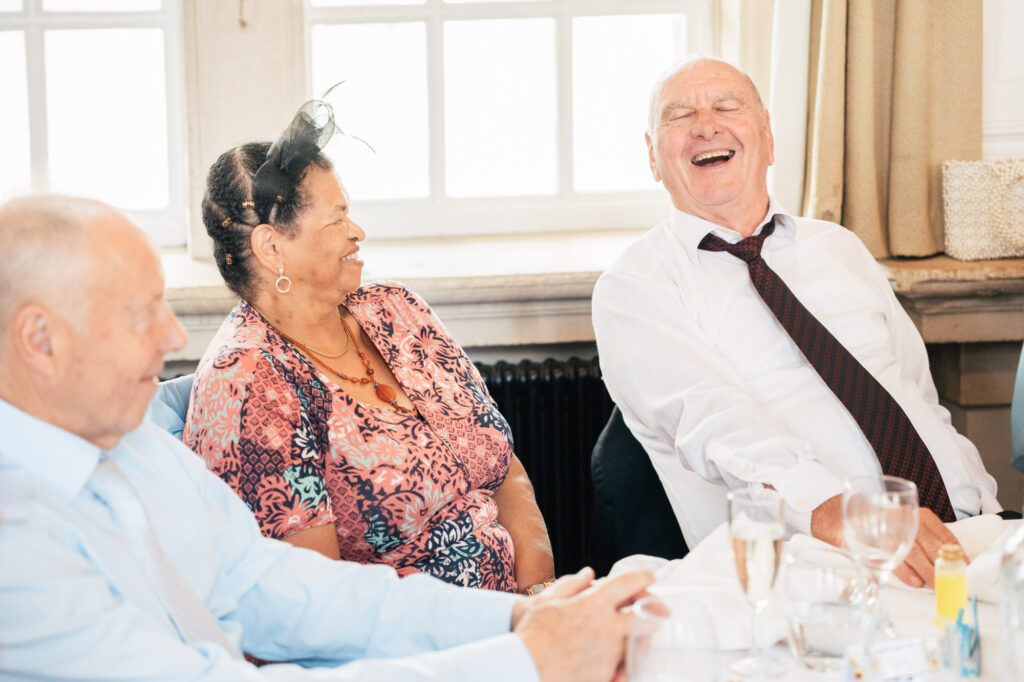 Three older adults sit at a table, smiling and laughing together in a bright room. The man on the right is leaning back, laughing heartily, while the others smile warmly. The table has glasses and plates.