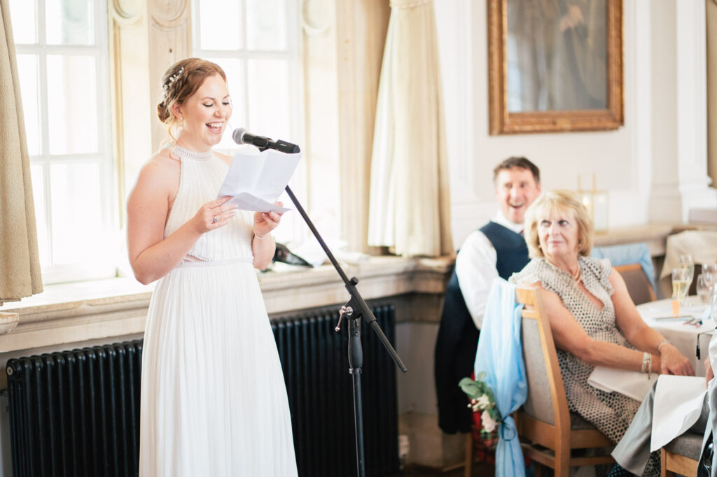 A woman in a white dress stands at a microphone, smiling as she reads from a paper at a bright indoor event. Seated guests listen attentively, and sunlight streams through large windows behind her.