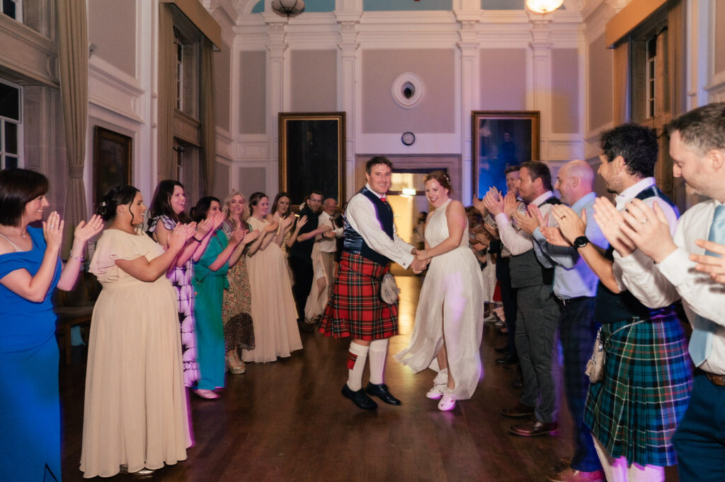 A bride and groom in traditional Scottish attire hold hands and smile while dancing. Guests form two lines, clapping and cheering inside a grand hall with high ceilings and large windows.