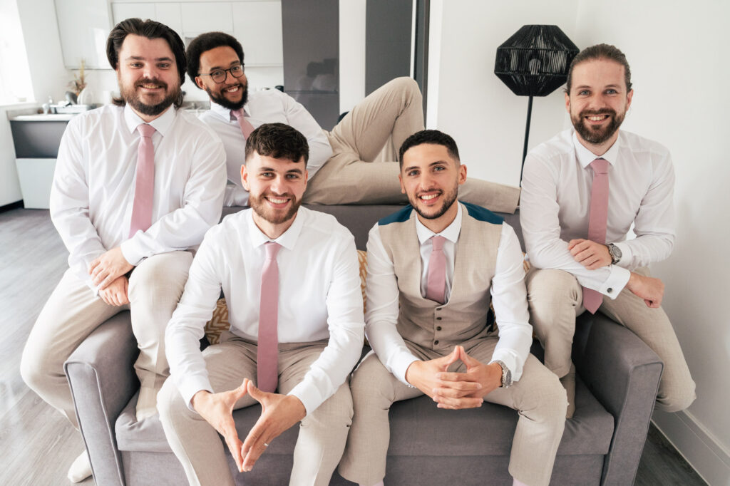 Five men dressed in light-colored suits and pink ties are smiling and sitting together on a sofa in a bright room, with one man lounging across the sofa’s backrest.