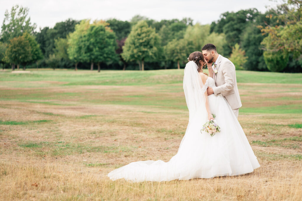 A bride and groom stand close together outdoors on a grassy field, sharing an intimate moment. The bride wears a long white dress and veil, holding a bouquet, while the groom is in a beige suit. Green trees are in the background.