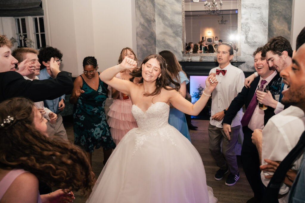 A bride in a white strapless gown dances joyfully, surrounded by smiling guests in formal attire at a wedding reception. Everyone appears to be having a great time on the dance floor.