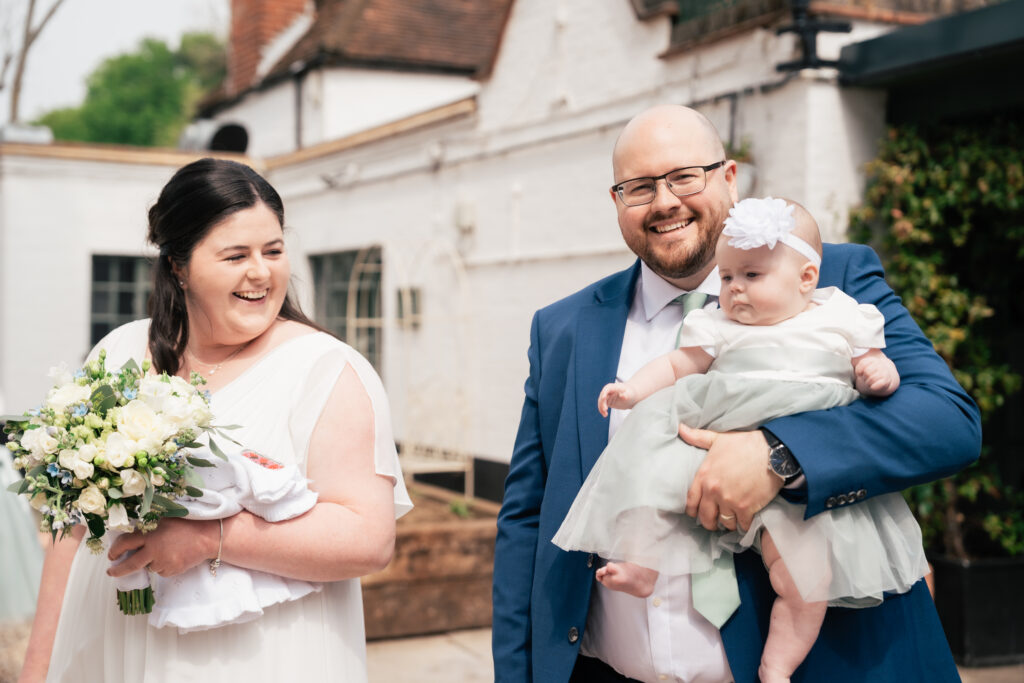 A smiling woman in a white dress holds a bouquet beside a man in a blue suit holding a baby in a white dress and headband. Captured outdoors near white buildings, this joyful moment was beautifully preserved by their Surrey wedding photographer.
