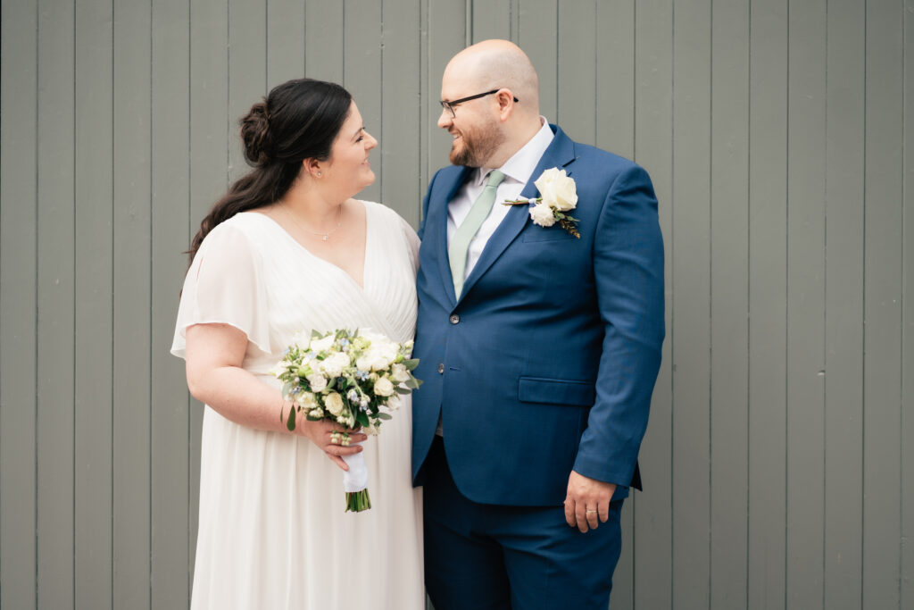 A bride in a white dress holding a bouquet and a groom in a blue suit with a white rose boutonniere stand together, smiling at each other, as their Surrey wedding photographer captures the moment by a gray wooden wall.