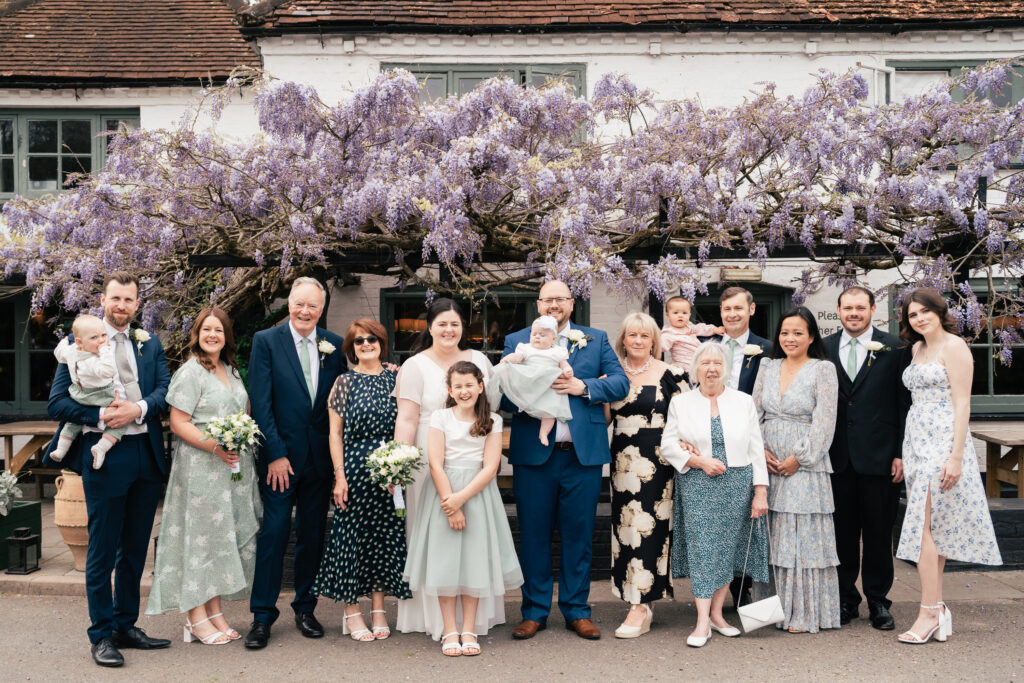 A large group of people, including adults and children, pose for a formal photo outside under blooming purple wisteria. Many wear formal or semi-formal attire, smiling in front of a white building with flowers—captured by a Surrey wedding photographer.