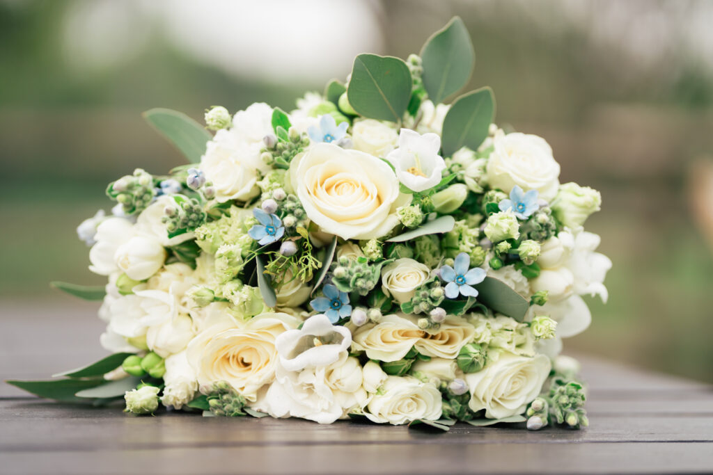 A bouquet of white and pale yellow roses, small blue flowers, and green leaves rests on a dark wooden surface outdoors—a perfect detail for any Surrey wedding photographer capturing natural elegance.