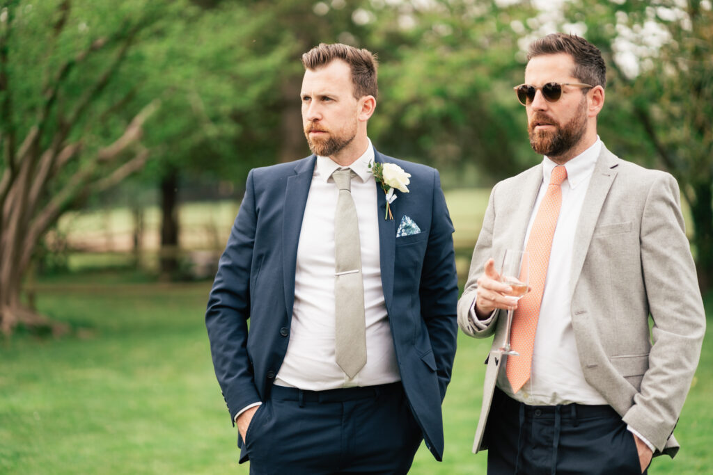 Two men dressed in suits stand outdoors on grass, captured by a Surrey wedding photographer. One wears a navy suit with boutonnière; the other sports a light gray jacket, orange tie, sunglasses, and holds a wine glass.