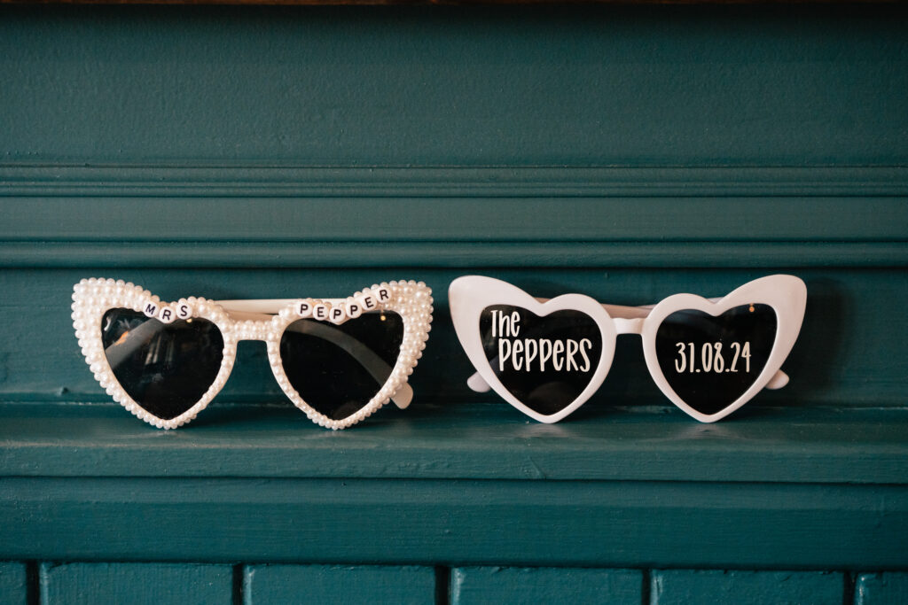 Two pairs of heart-shaped sunglasses with white frames rest on a teal shelf; one says Mrs Pepper with pearl decoration, the other says The Peppers and 31.08.24 in white text—a perfect detail for any Surrey wedding photographer to capture.