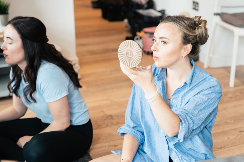A woman in a blue shirt holds a round compact mirror while applying makeup. Another woman in a light blue top sits beside her. Both are seated on a wooden floor in a bright room with bags and a chair in the background.