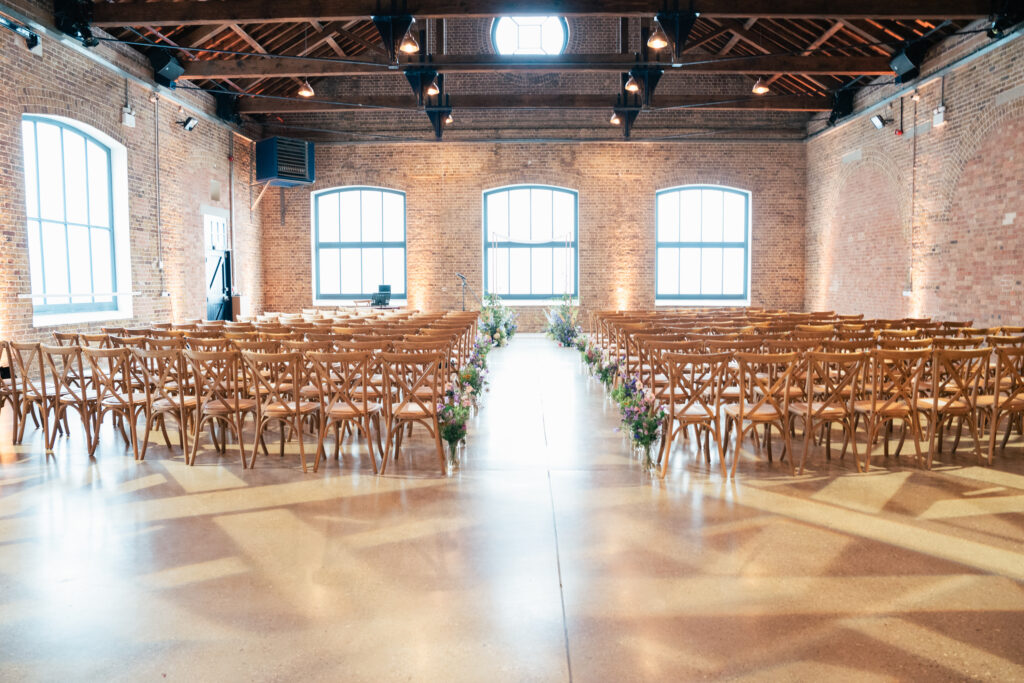 A spacious indoor wedding venue with rows of wooden chairs facing forward, floral arrangements along the aisle, large arched windows, brick walls, and exposed wooden beams on the ceiling.