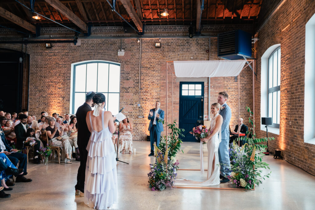 A couple stands under a modern chuppah during their indoor wedding ceremony, surrounded by flowers. Guests are seated, and a woman in a white ruffled dress reads from a paper as the officiant stands nearby. The venue features brick walls and large windows.