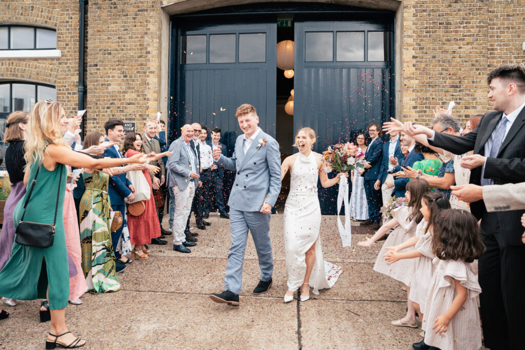 A newlywed couple, smiling and holding hands, walks outside a brick building while guests standing on either side cheer and throw confetti. The bride holds a bouquet and both are dressed in light-colored outfits.