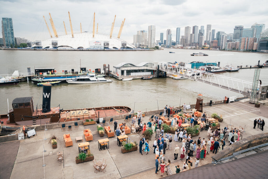 A large group of people gathers at an outdoor riverside venue with seating and plants. Boats are docked nearby, and a city skyline with tall buildings and the O2 Arena is visible across the water under a cloudy sky.