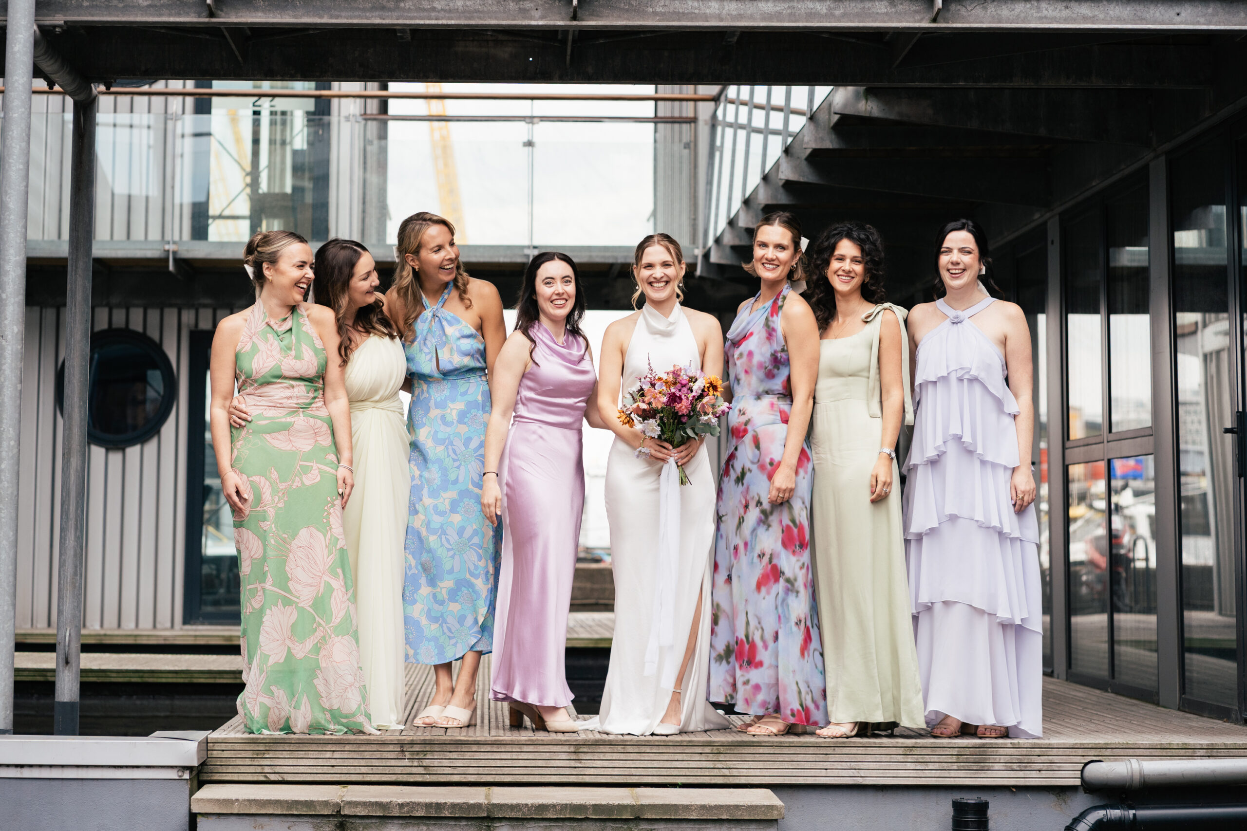 A bride in a white dress stands with seven bridesmaids in colorful dresses, all smiling on wooden steps outside a modern building with glass and metal features. The bride holds a bouquet of flowers.