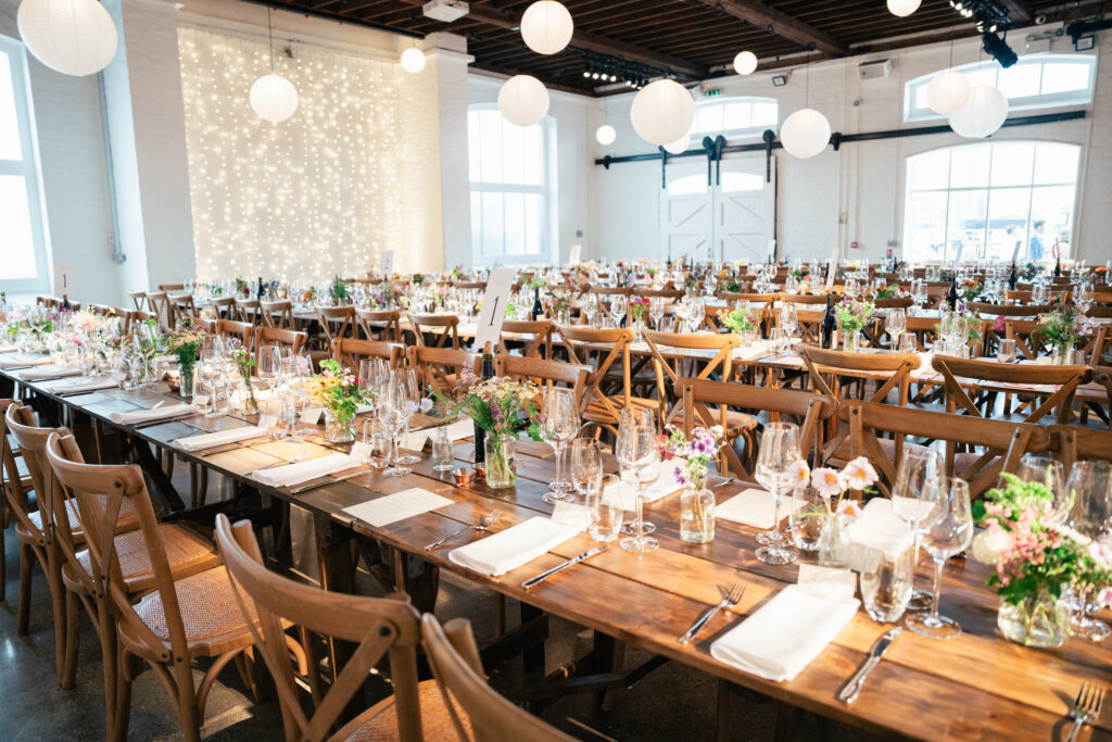 A spacious event hall with long wooden tables set for a formal meal, featuring floral centerpieces, glassware, and napkins. White paper lanterns and string lights hang from the ceiling, creating a bright, festive atmosphere.