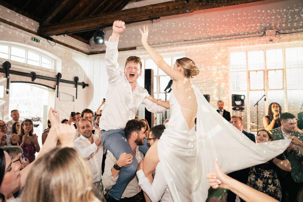 A bride and groom are lifted on chairs by friends at a lively wedding reception, surrounded by cheering guests under sparkling lights in a bright, rustic venue.