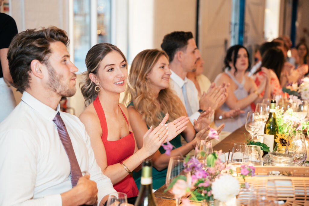 People dressed in semi-formal attire sit at a long table decorated with flowers and wine, smiling and clapping at an event or celebration. The atmosphere appears joyful and lively.