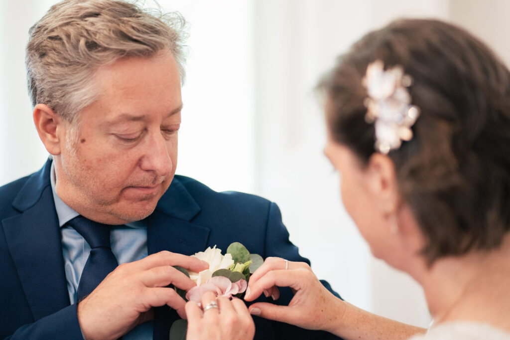A woman pins a boutonniere with white and pink flowers onto a man’s dark suit jacket. The man looks down at the flowers, and the woman’s hair is adorned with a decorative hair clip. Both appear focused and calm.