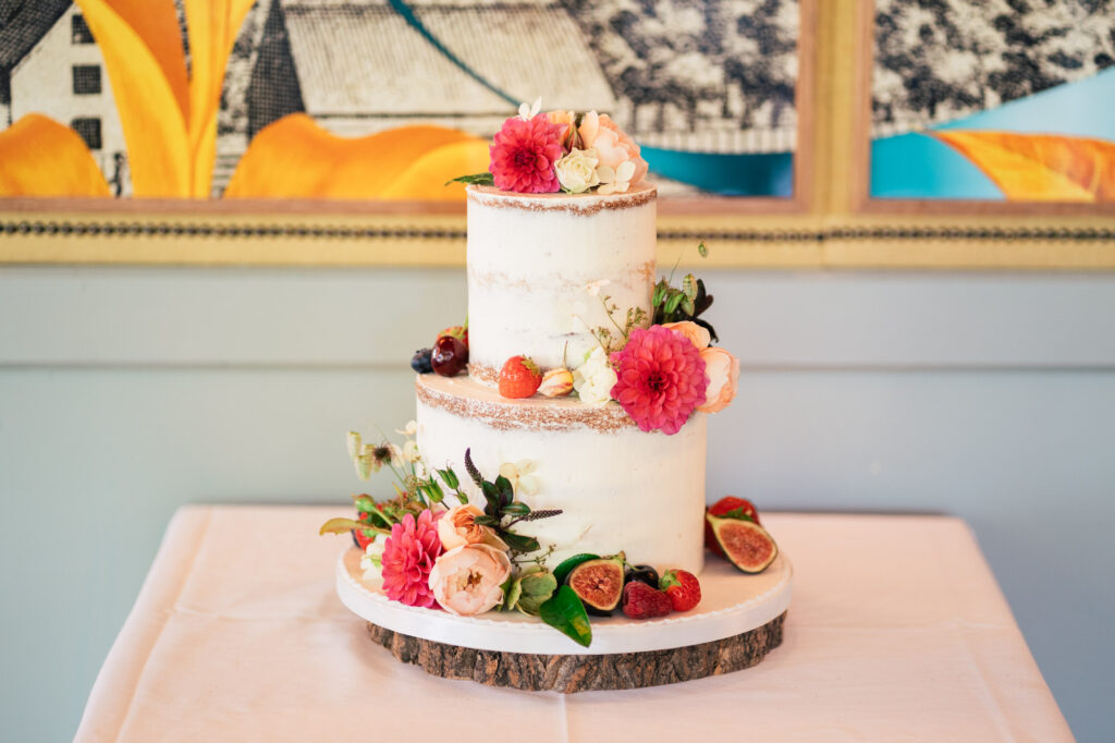A two-tiered semi-naked cake decorated with pink and white flowers, strawberries, figs, and greenery sits on a wooden cake stand atop a white tablecloth. A colorful mural is visible in the background.
