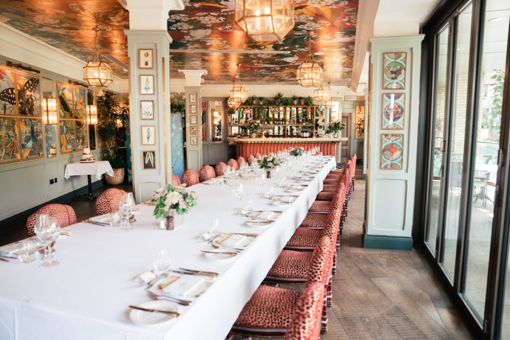 A long dining table with white tablecloth, set with plates, glasses, and flower centerpieces, sits in an elegant restaurant with red chairs, colorful ceiling art, large windows, and a bar in the background.