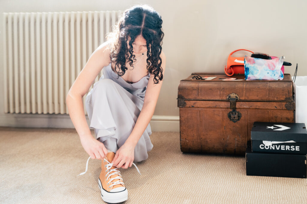A woman in a lavender dress kneels on a beige carpet, tying the laces of tan Converse sneakers—a candid scene a Surrey wedding photographer would love. Beside her are a wooden chest, colorful makeup bag, and black Converse shoe boxes.