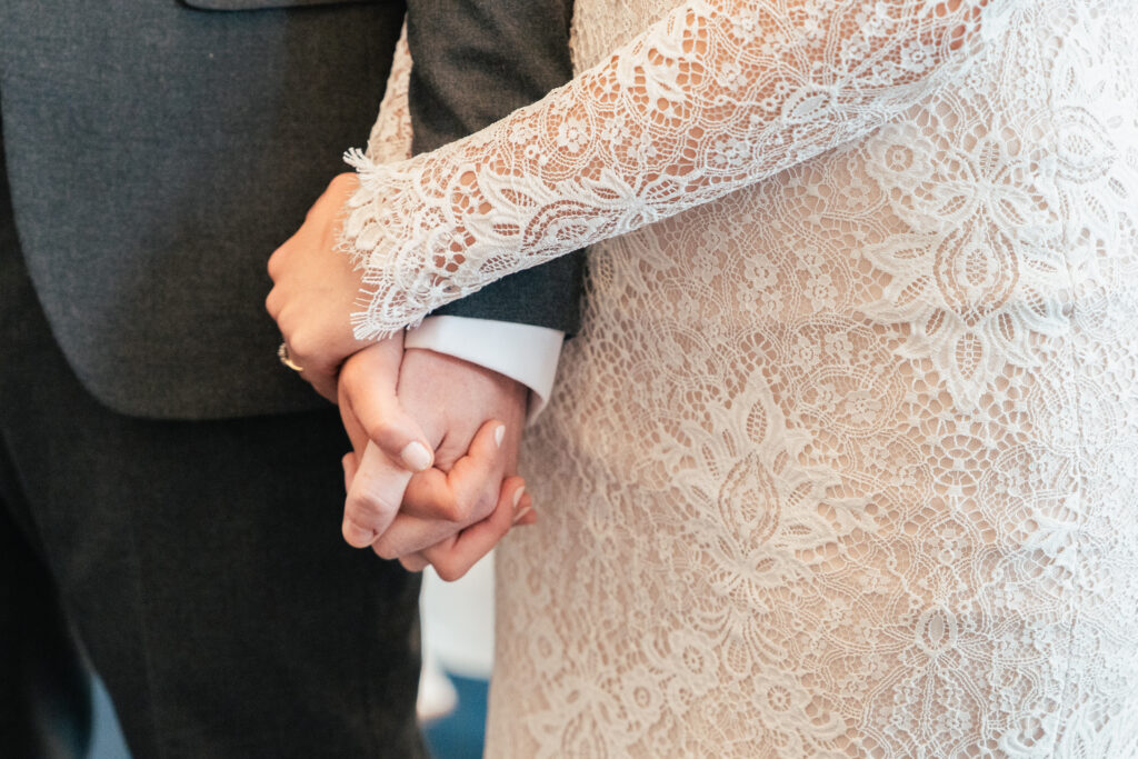 Close-up of a couple holding hands, with one person in a lace wedding dress and the other in a dark suit—captured by a Guildford Registry Office photographer.