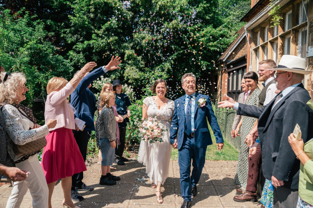 A newlywed couple smiles as they walk outside the Guildford Registry Office, surrounded by cheering guests throwing confetti. The bride holds a bouquet in her white dress, while the groom wears a suit. It’s a sunny day with trees and buildings in the background.