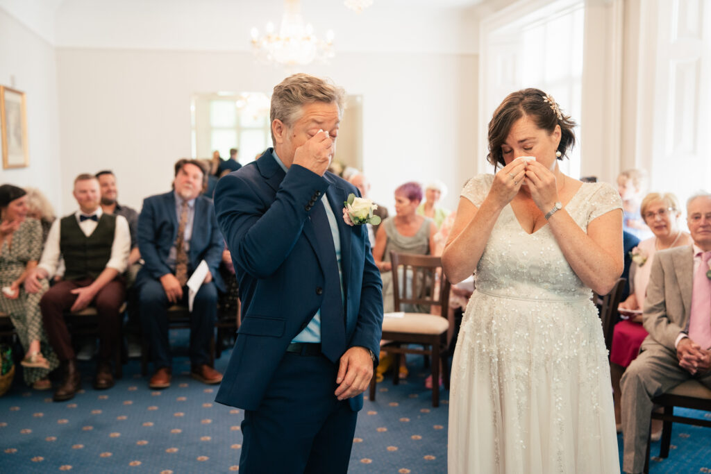 A bride and groom stand at the front of a room, both wiping away tears, while guests watch the emotional moment. Captured by a Guildford Registry Office photographer, this scene beautifully reflects the heartfelt emotions of their wedding ceremony.