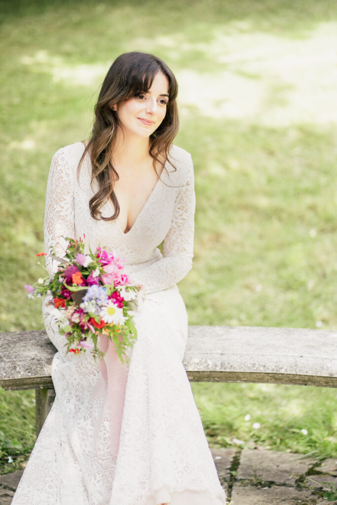 A woman in a white lace wedding dress sits on a stone bench outdoors, holding a colorful bouquet and smiling softly—captured beautifully by a Guildford Registry Office photographer. Green grass fills the blurred background.