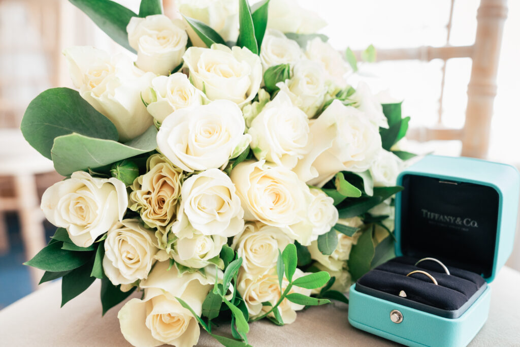 A bouquet of pale yellow roses with green leaves sits next to a Tiffany & Co. ring box containing two gold rings on a table, beautifully captured by a Guildford Registry Office photographer.