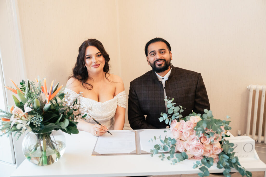 A bride in an off-shoulder white dress and a groom in a dark suit smile while signing a document, captured by a Guildford Registry Office photographer. The table is adorned with pink roses and a vase of flowers.