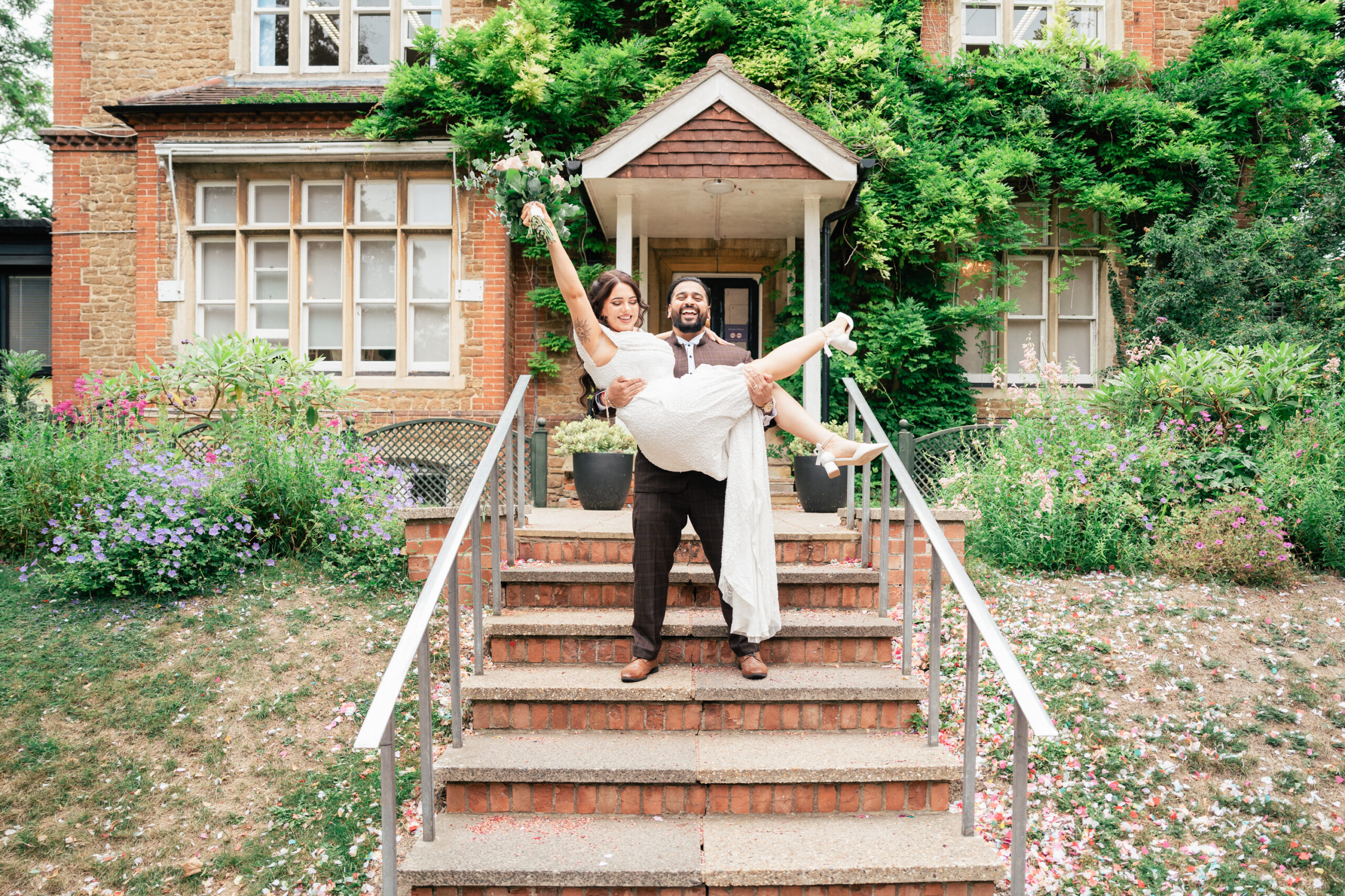 A joyful groom holds his smiling bride in his arms on steps outside a brick house, surrounded by greenery and flowering plants. Captured by a Guildford Registry Office photographer, the bride lifts her bouquet and leg in celebration.